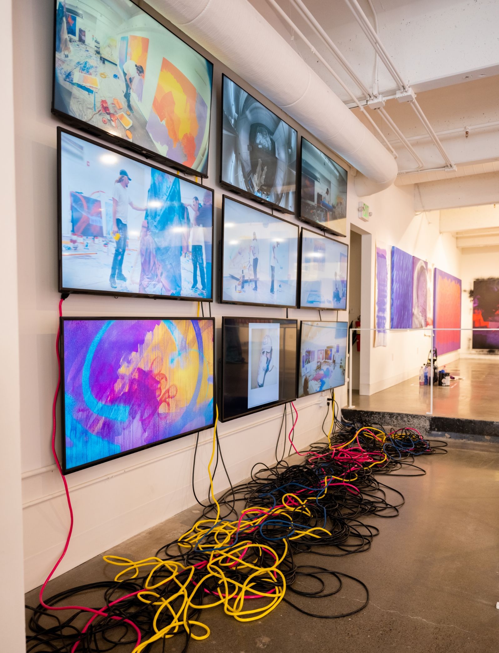 Wall with nine video screens in a grid showing behind the scenes footage of the artists creating the art in show. Large pile of cords sit on floor below screens