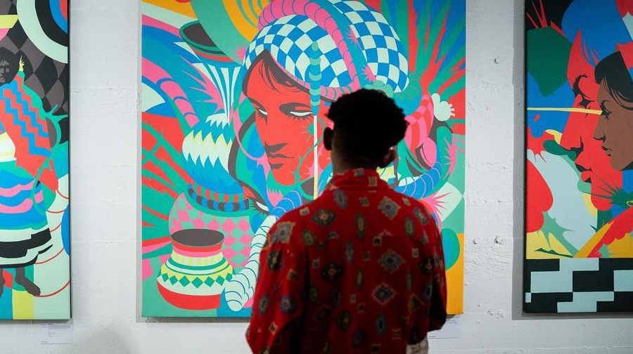 A person in a red patterned shirt views colorful, abstract paintings displayed on a white wall in a gallery.