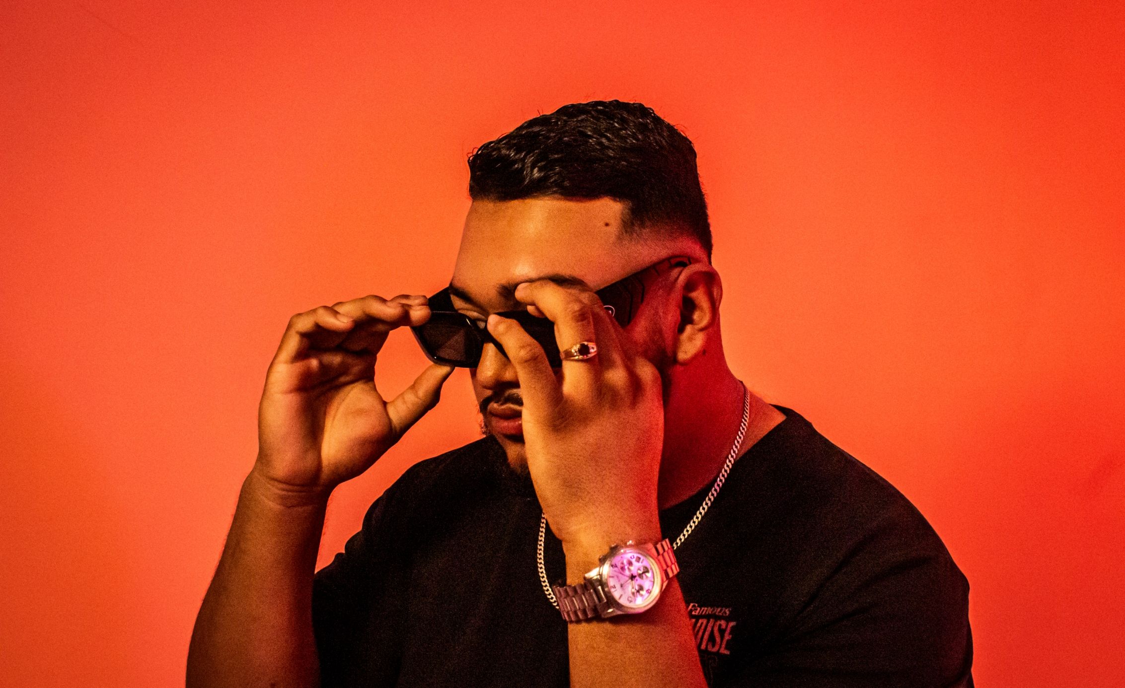 A man adjusting his sunglasses against a vibrant red background. He wears a watch and a chain, and has a neatly styled haircut.