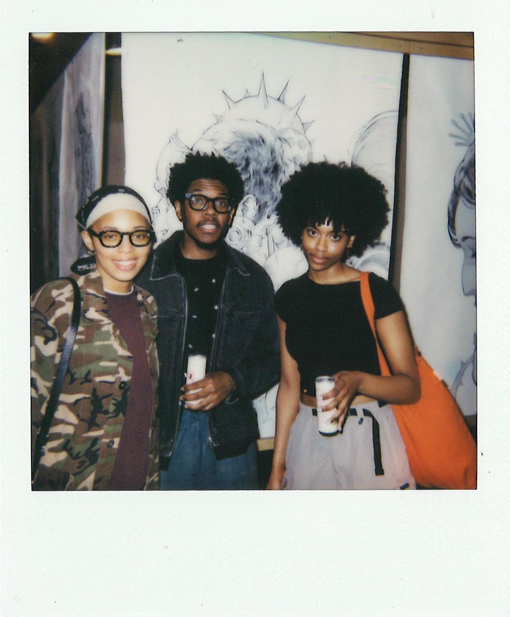 Three people stand together in front of artwork, holding drinks. One person wears a camouflage jacket, another a denim jacket, and the third wears a black shirt and orange bag.