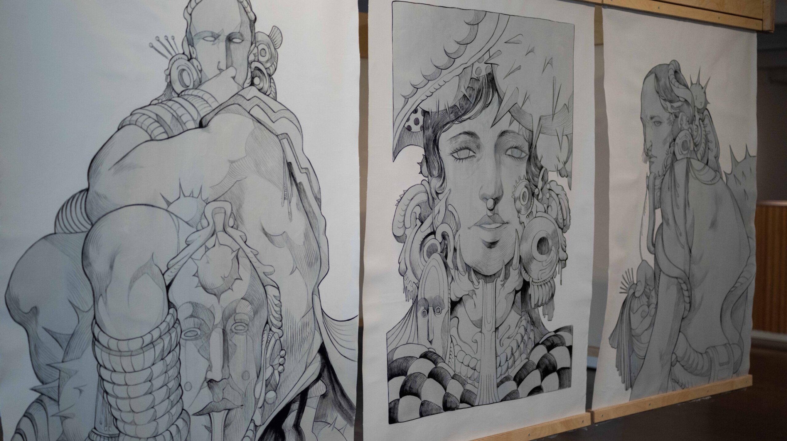 Three large pencil drawings on paper featuring abstract human figures with intricate, ornate designs are displayed side by side on wooden frames.