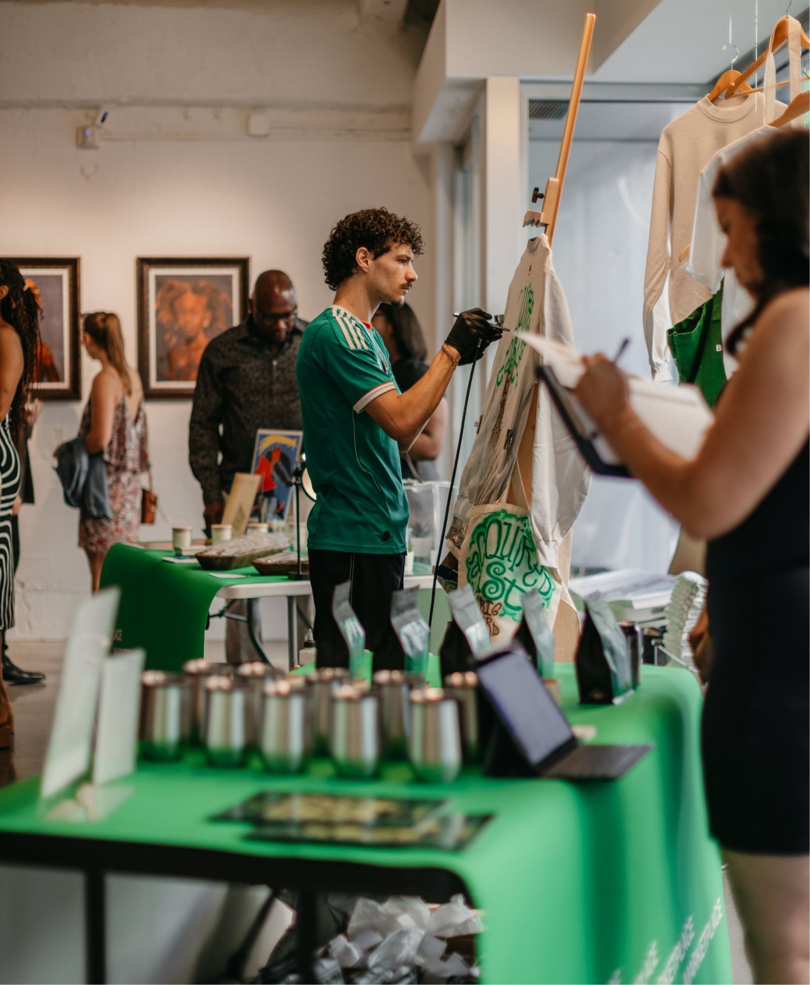 A man paints on a canvas at an indoor art event. Several people browse nearby tables displaying artwork and merchandise.