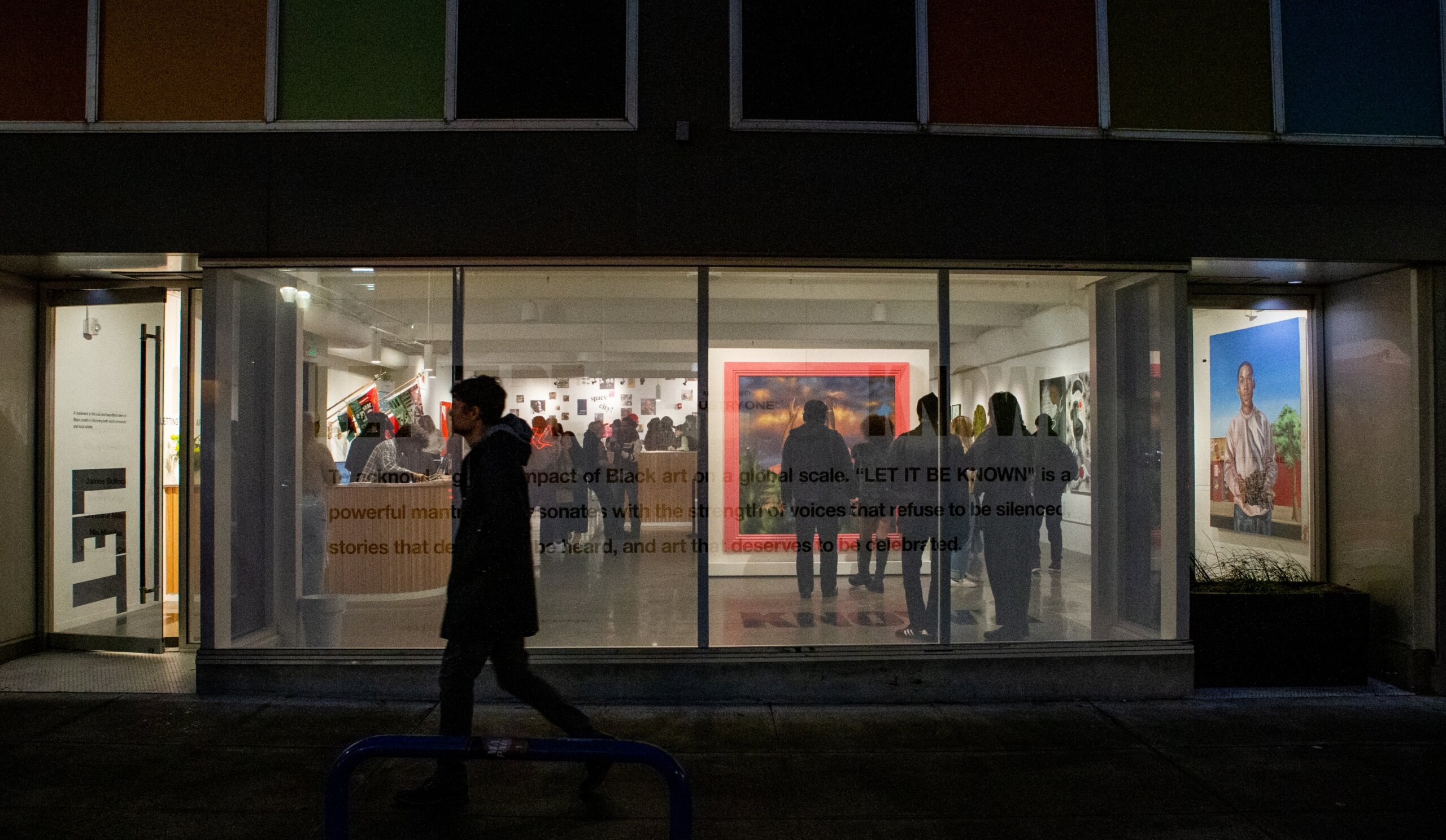 night time phonto of Industry One art Gallery front window. A person walks by on sidewalk and the inside of gallery is lit up showing many people and art pieces inside the gallery