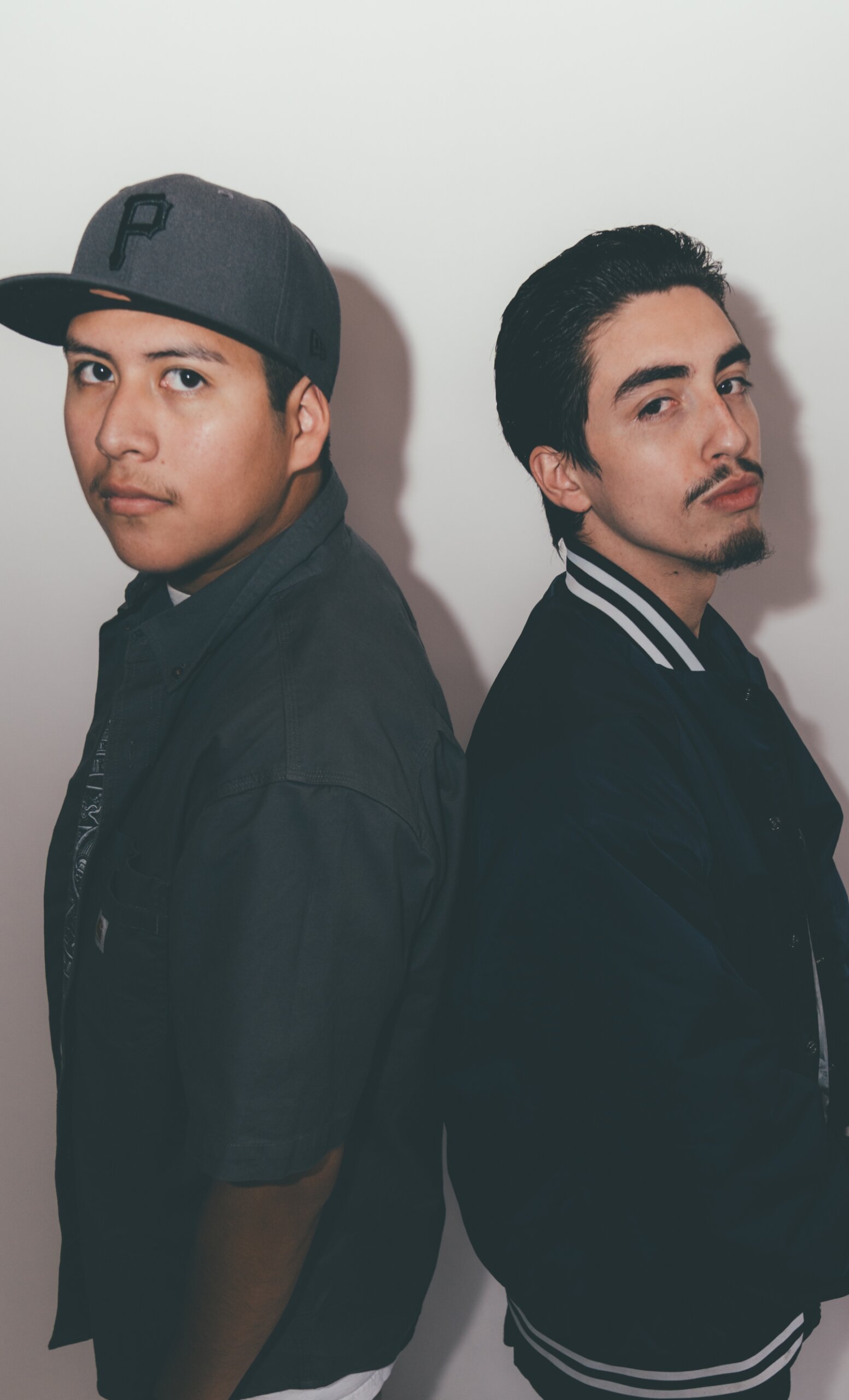 Two young men posing back-to-back, one wearing a gray cap and shirt, and the other in a dark varsity jacket.