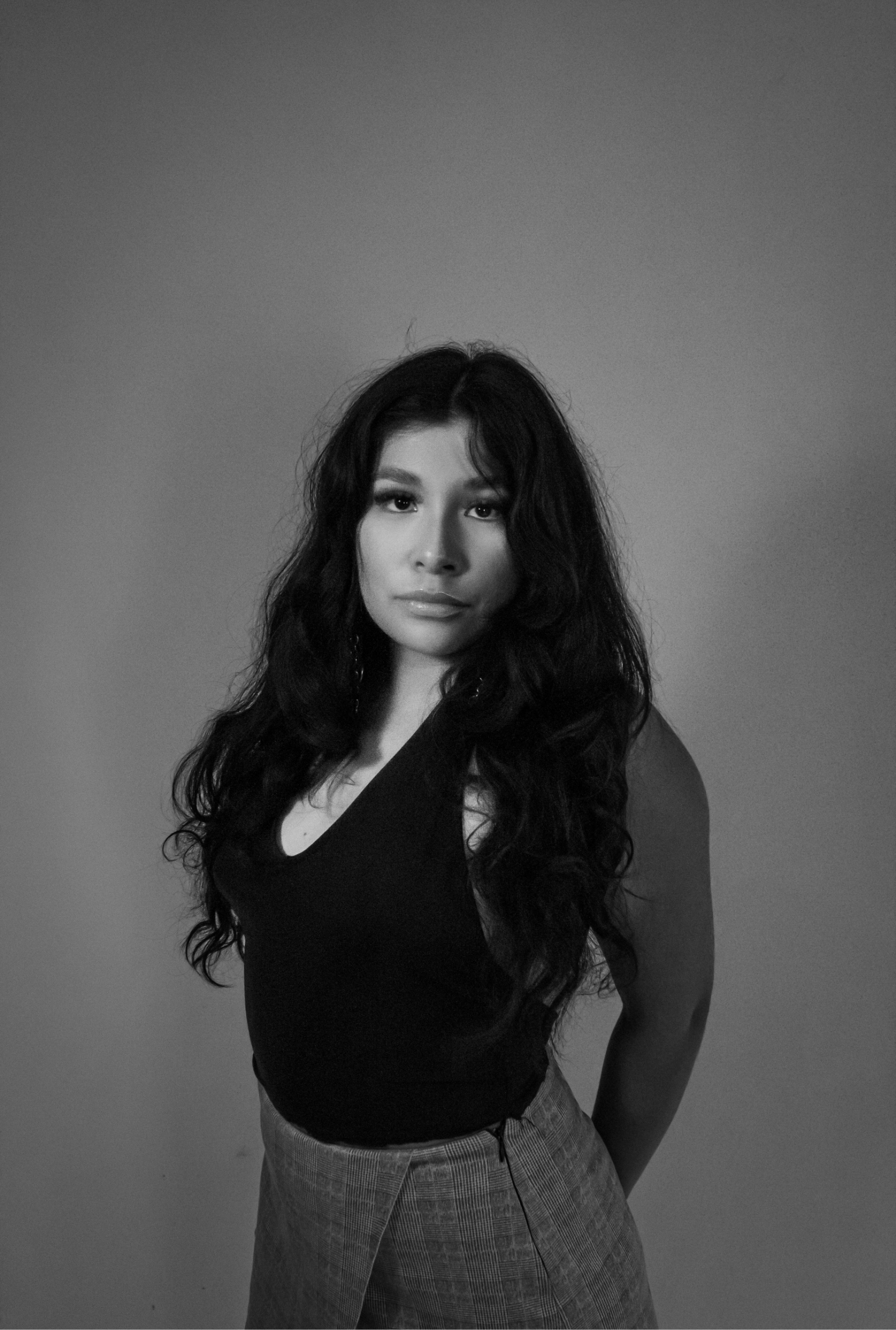 Black and white portrait of a woman with long curly hair, wearing a sleeveless top and plaid trousers, standing against a plain background.