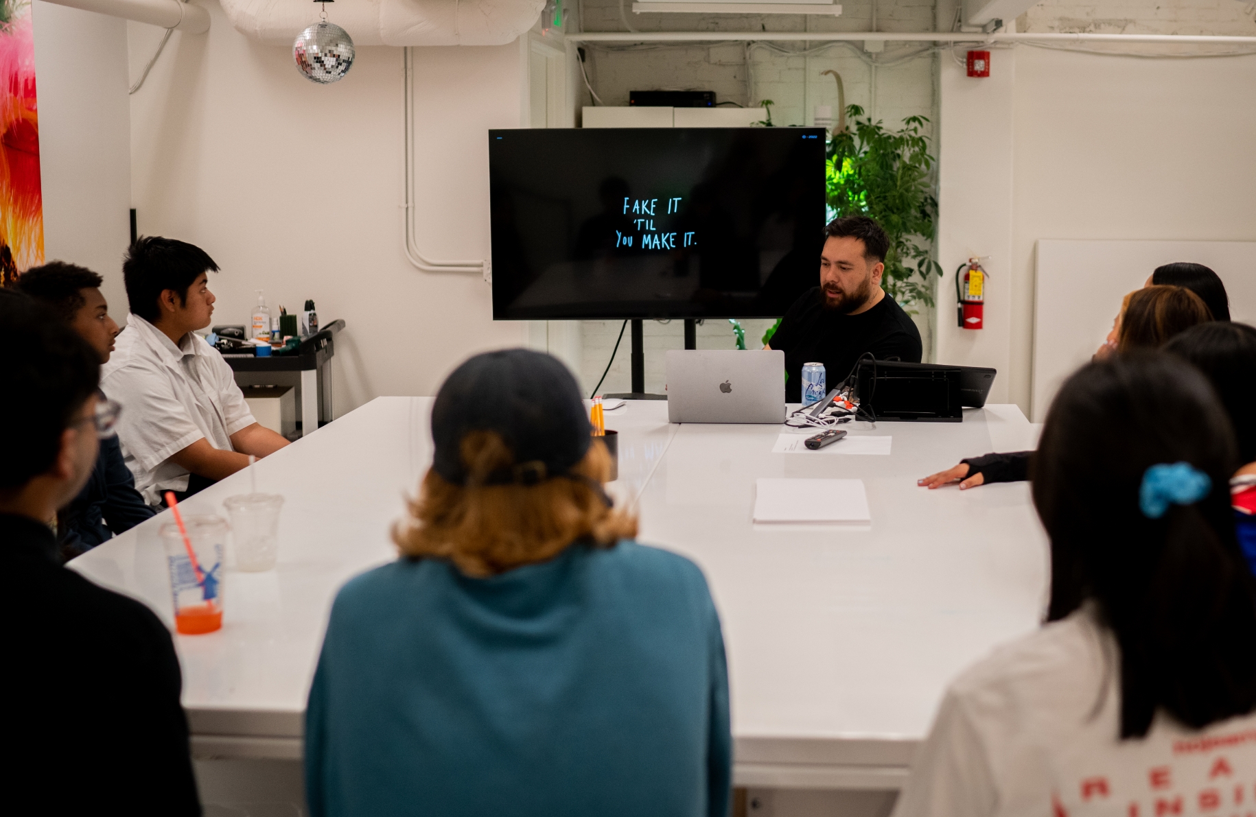 A diverse group of individuals sitting at a conference table attentively listening to a presenter at a laptop with the phrase "Fake it 'til you make it" on screen.