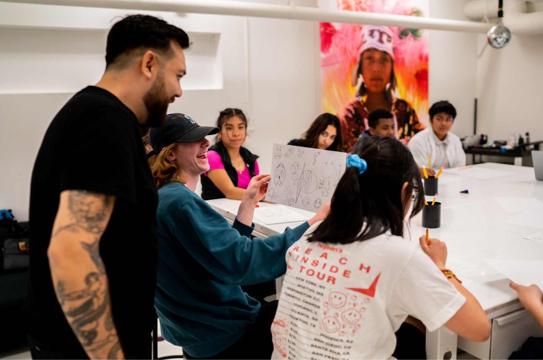 A diverse group of students in a classroom, interacting with their teacher who is reviewing a student's artwork.