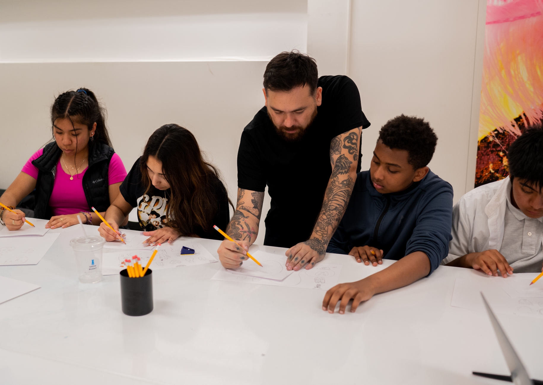 A diverse group of students sitting at a table, engaged in drawing, with an adult male overseeing their work in an art classroom.