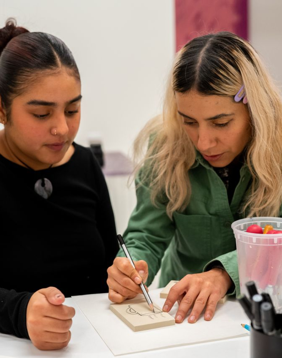 Two people sitting at a table, one writing on a notepad, the other observing. A container with colorful items is on the table.