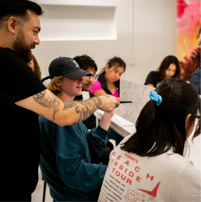 A group of people in a classroom setting; an instructor with tattoos gestures towards a sketch, while students observe and take notes.