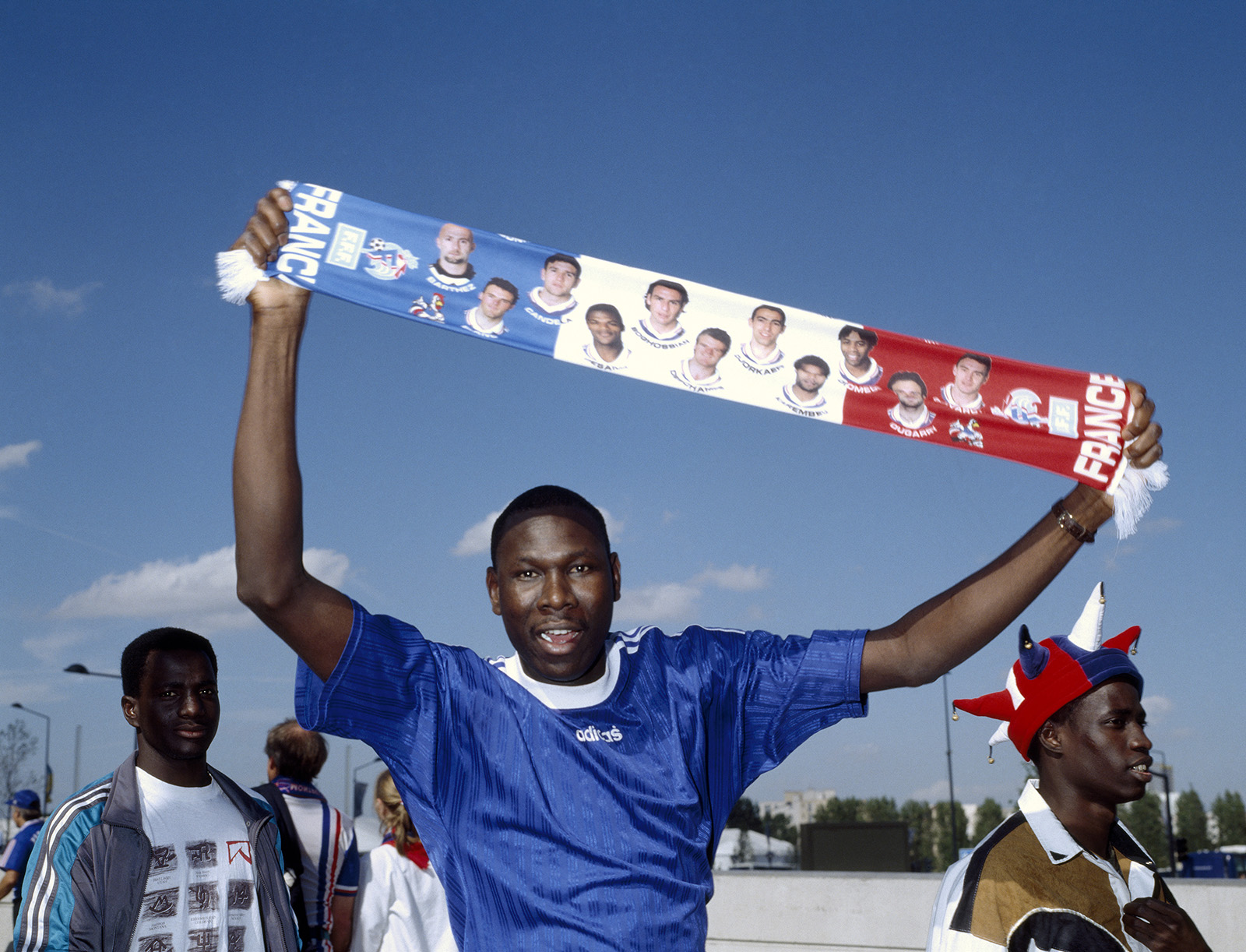 A man in a blue France football jersey holds up a scarf featuring players' photos, with "FRANCE" written on both ends, while others stand nearby under a clear sky.